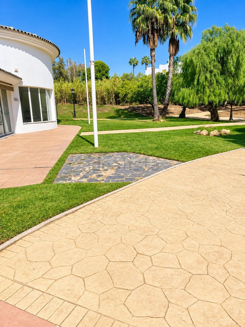 A paved patio area leads to a lush green lawn with palm trees and lush foliage under a clear blue sky.