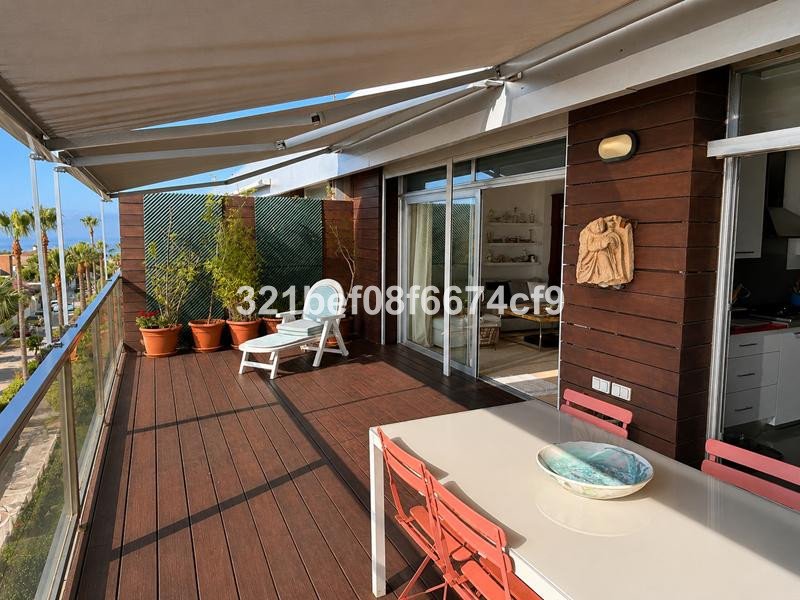 Spacious wooden balcony with outdoor dining set, lounge chair, plants, and a partial view of palm trees and the sea.