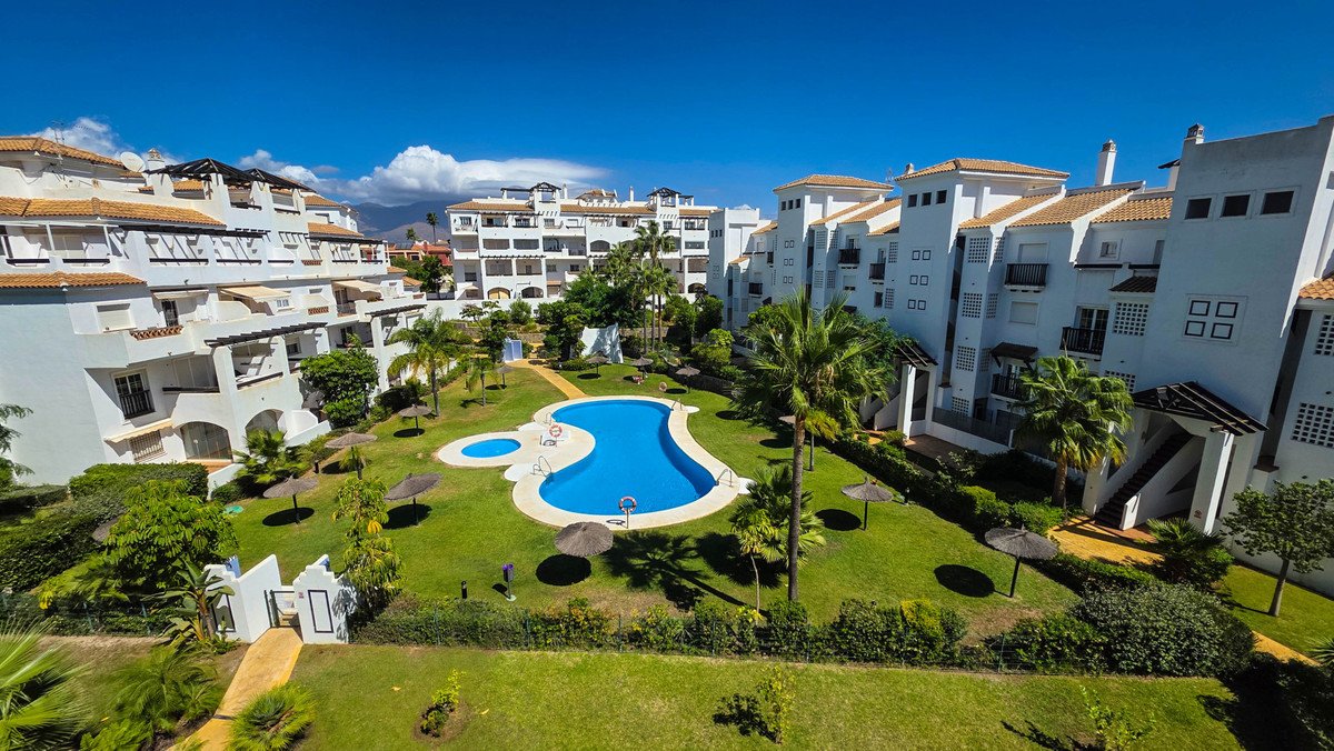 A view of apartments surrounding a resort-style swimming pool, with lush green lawns and palm trees under a clear blue sky.