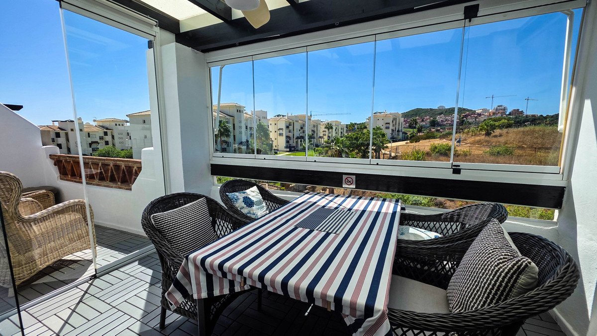 A covered terrace with seating and a table, offering a view of apartment buildings and a hillside under a clear blue sky.