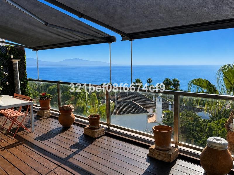 A wooden terrace with outdoor seating and potted plants, offering a stunning view of the blue sea and distant mountains.