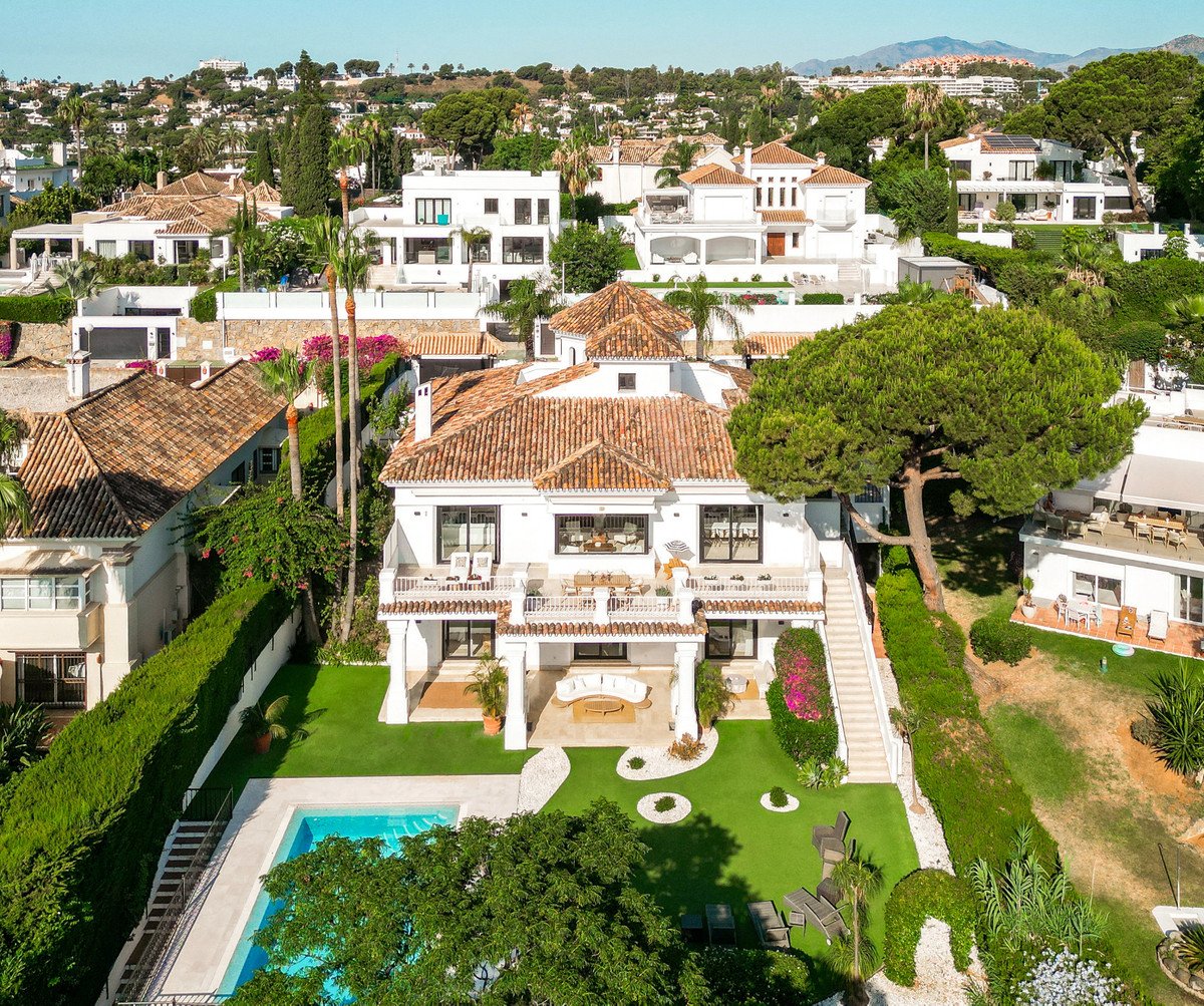 Aerial view of a luxurious white villa with terracotta roof, pool, and lush green garden in a residential area.