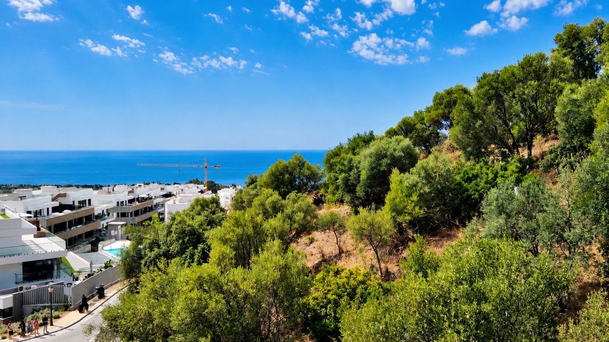 View of modern apartments with a pool, overlooking the sea and surrounded by lush green trees and hills.