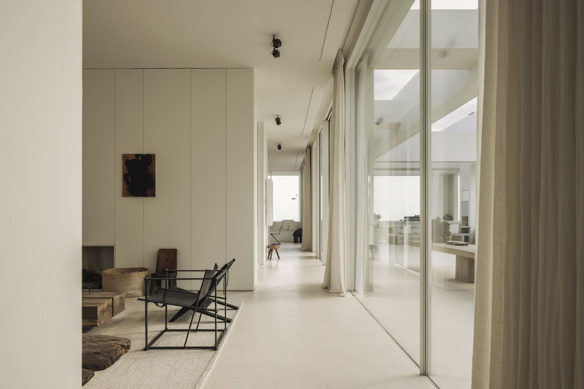 Modern minimalist living room with floor-to-ceiling windows, contemporary furniture, and artwork, overlooking the sea.