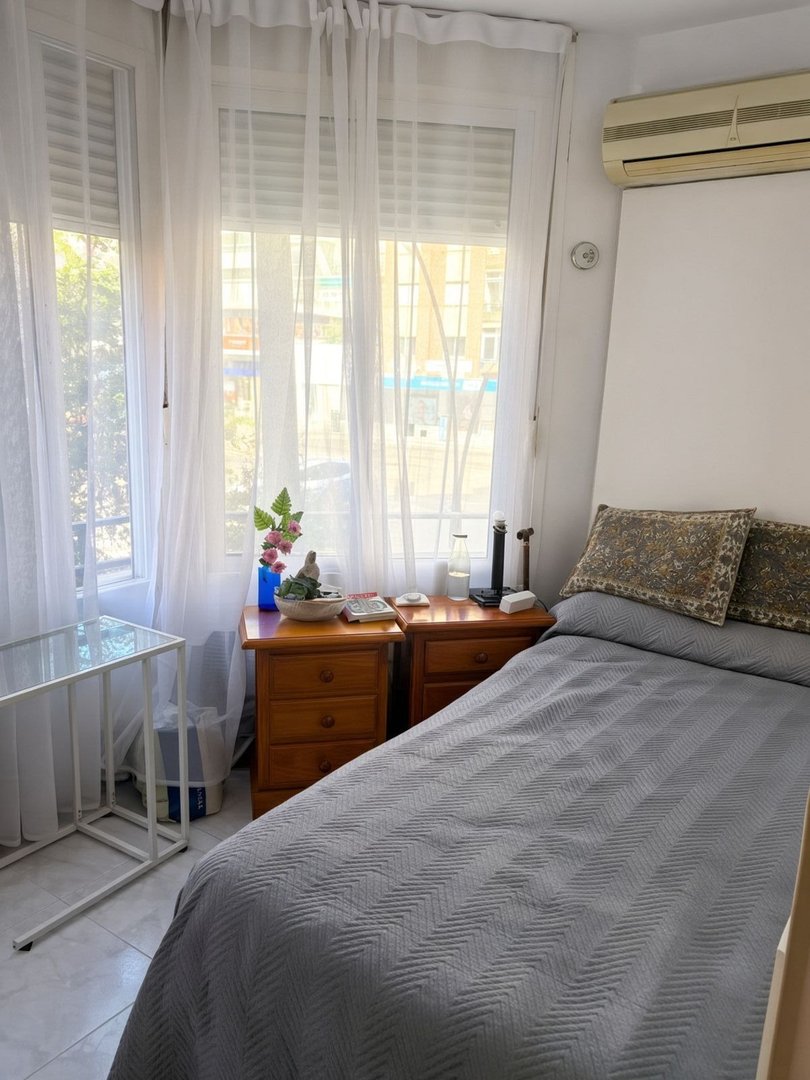 Cozy bedroom featuring a bed with grey bedding, bedside tables, and a large window with sheer curtains offering a view of the city.