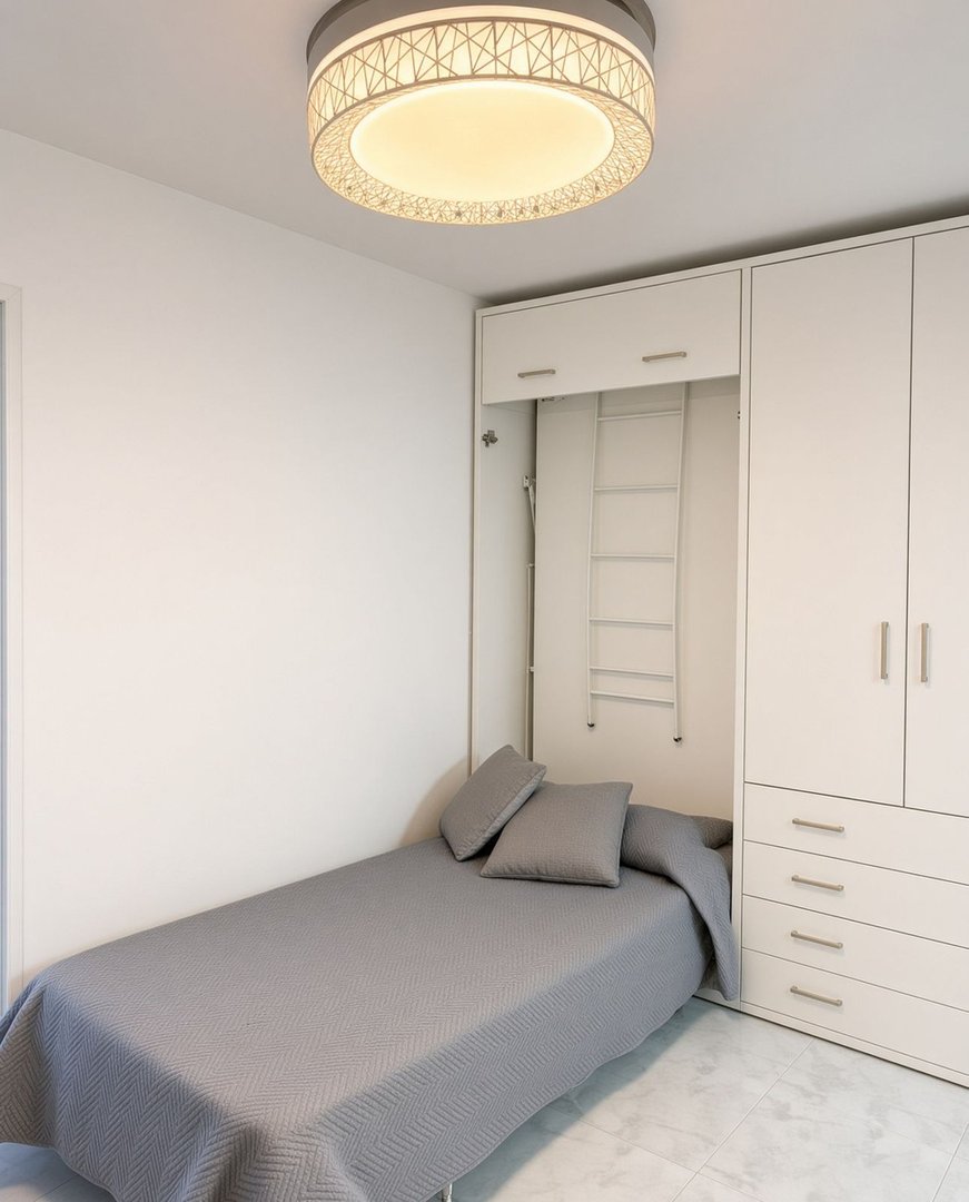 A compact bedroom with a murphy bed, grey bedding, pillows, and built-in white wardrobes with storage drawers.