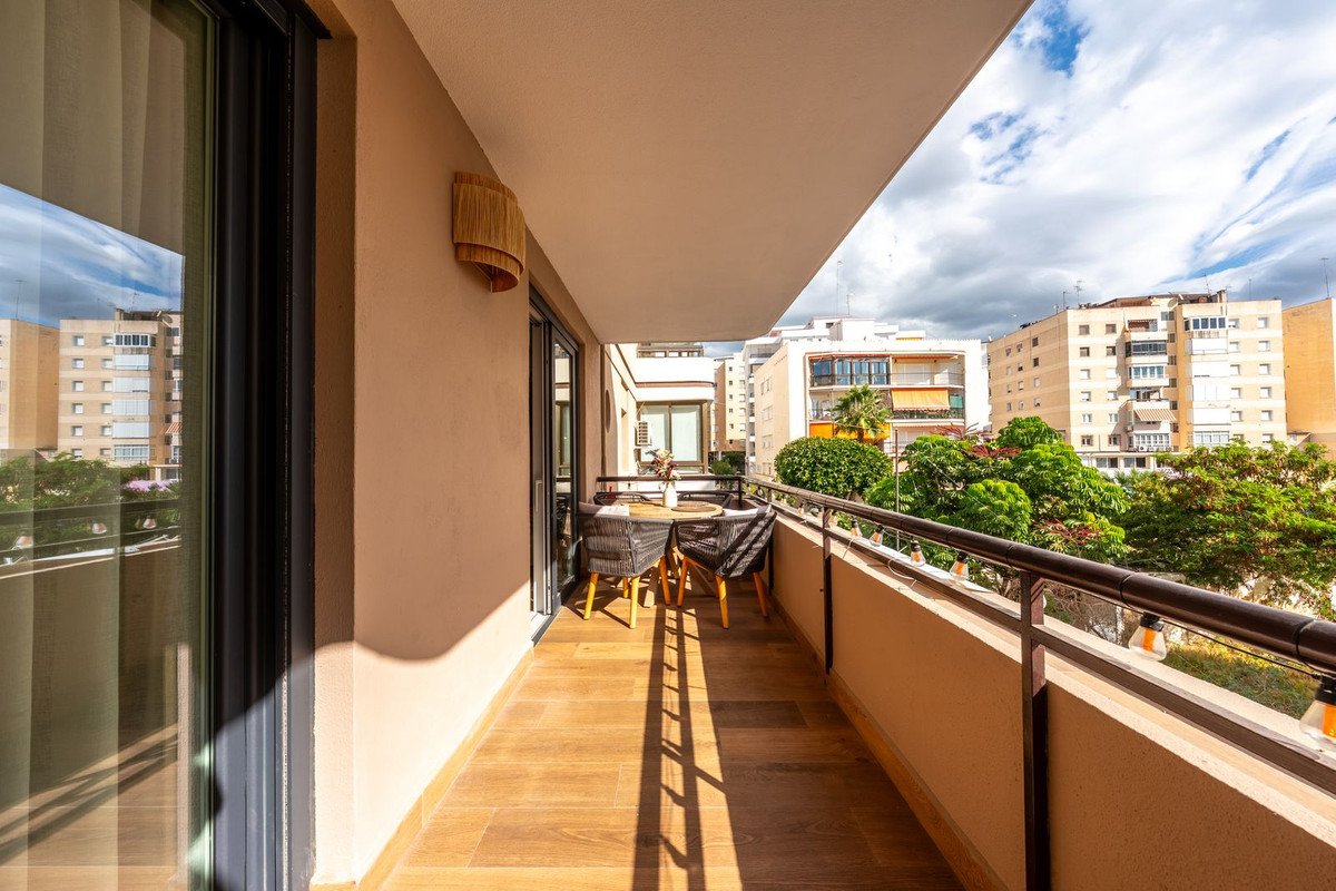 A spacious balcony with outdoor seating and a view of city buildings and green trees under a cloudy sky.