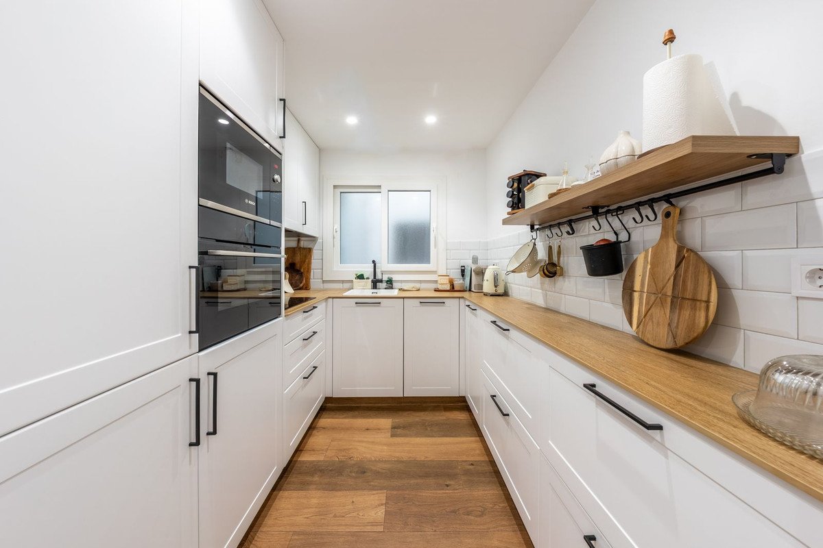 A modern white galley kitchen with wooden countertops, integrated appliances, and a small window above the sink.