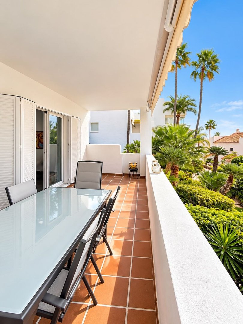 Terrace with a dining table and chairs overlooking lush greenery and palm trees under a blue sky.