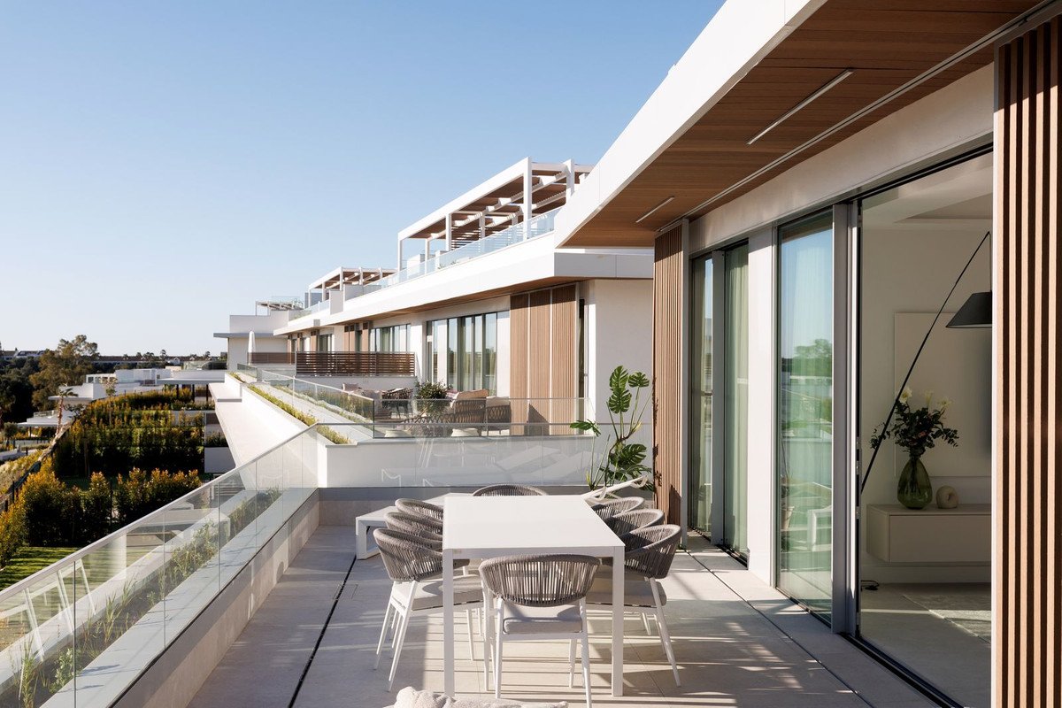Spacious modern terrace with dining table and chairs, offering a view of surrounding buildings and greenery under a clear blue sky.
