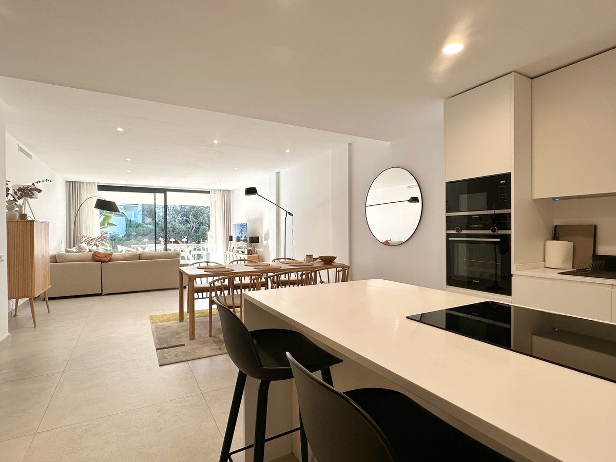 Modern open-plan living area with a kitchen island, dining table, and lounge seating, featuring large windows and a garden view.