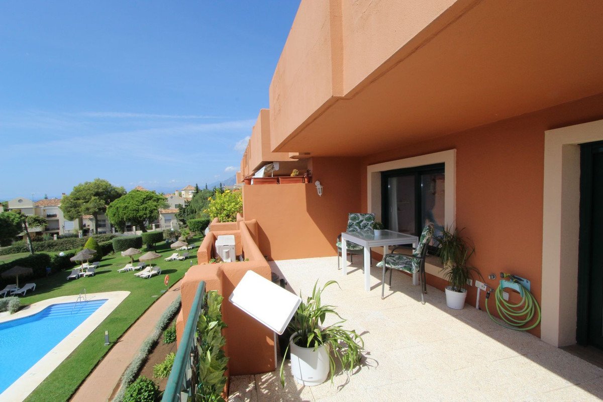 A spacious balcony with a table and chairs overlooks a communal pool and well-maintained gardens on a sunny day.