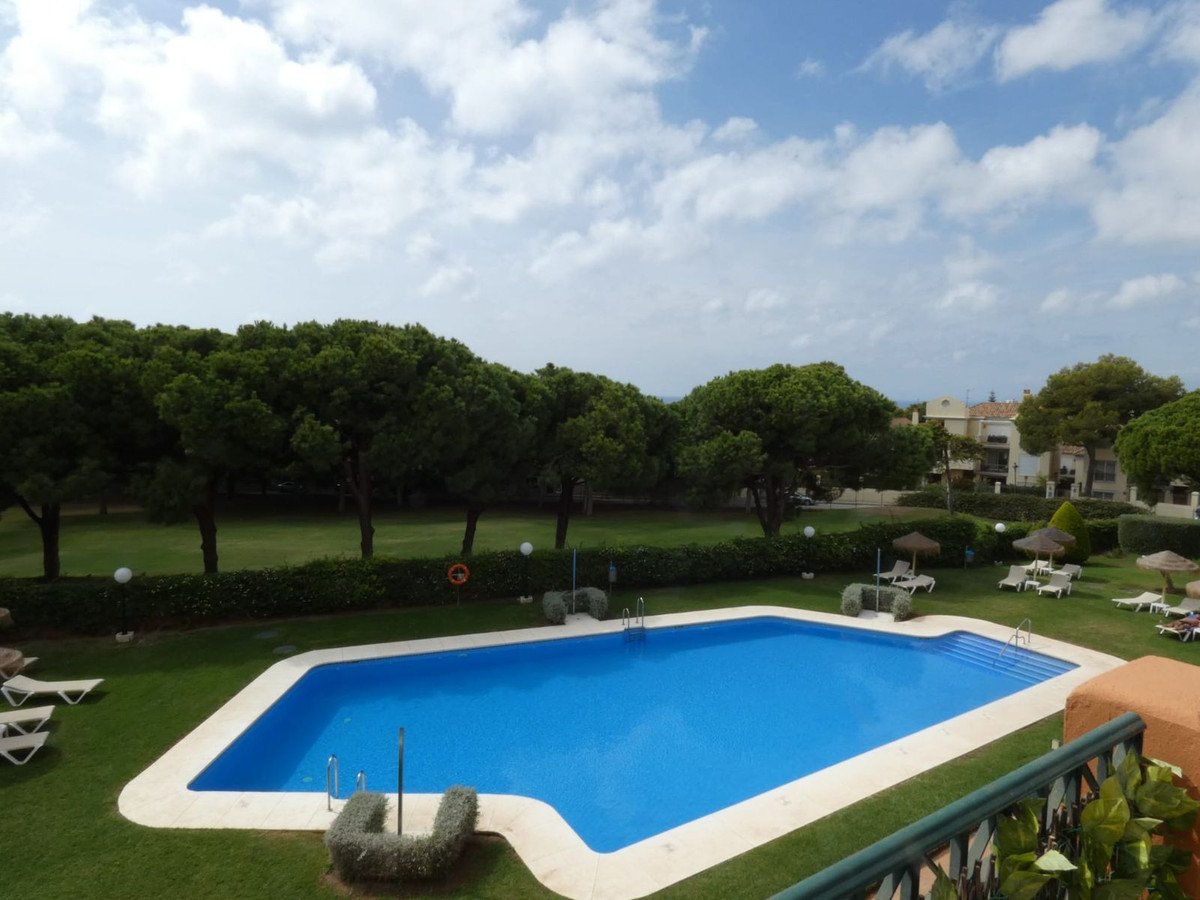 A blue swimming pool surrounded by a green lawn, pine trees, and apartment buildings under a cloudy sky.