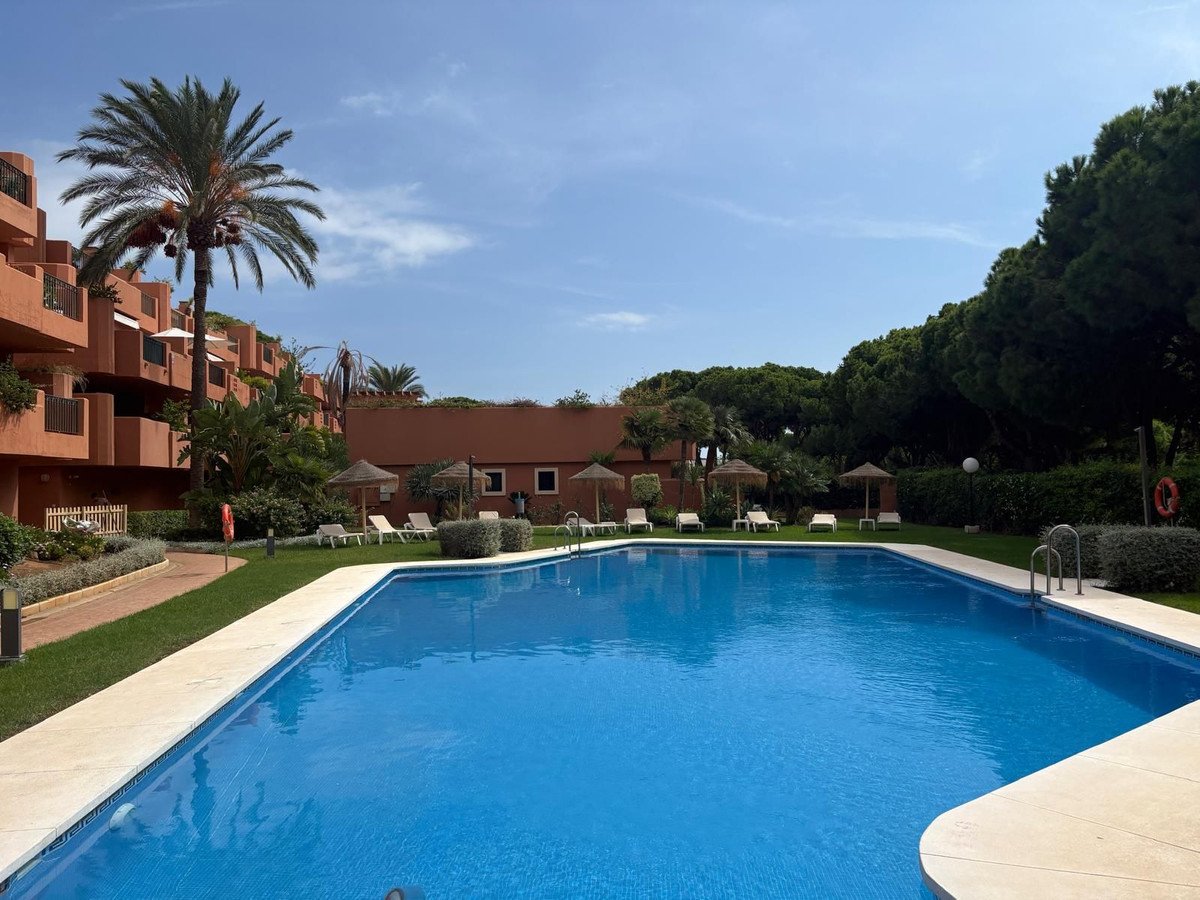 A blue swimming pool surrounded by green grass, lounge chairs, umbrellas, and a palm tree, with apartment buildings in the background.