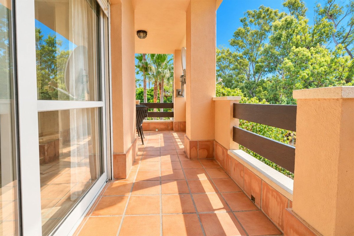 A sunny terracotta tiled balcony with a glass door, wooden railing, and lush green trees visible beyond.