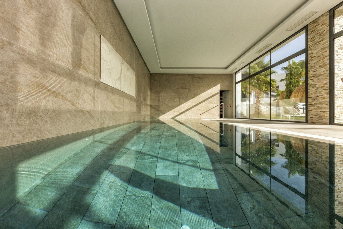 Luxurious indoor swimming pool with natural light streaming through large windows, offering a view of palm trees and outdoor lounge area.
