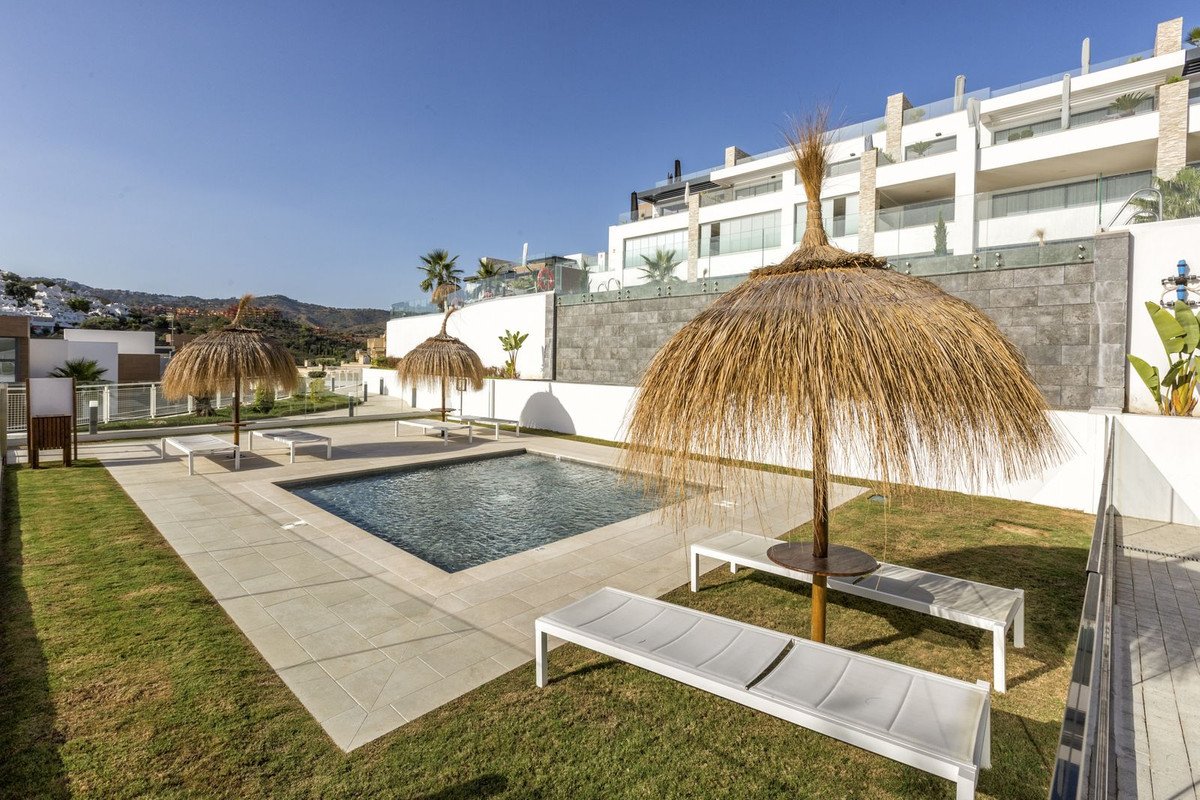 An outdoor swimming pool area with thatched umbrellas, sun loungers, and a modern building in the background under a clear blue sky.