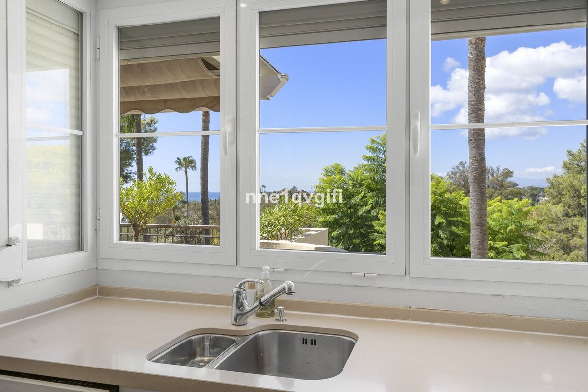 Kitchen sink with a view of palm trees, lush greenery, and the sea on a sunny day.