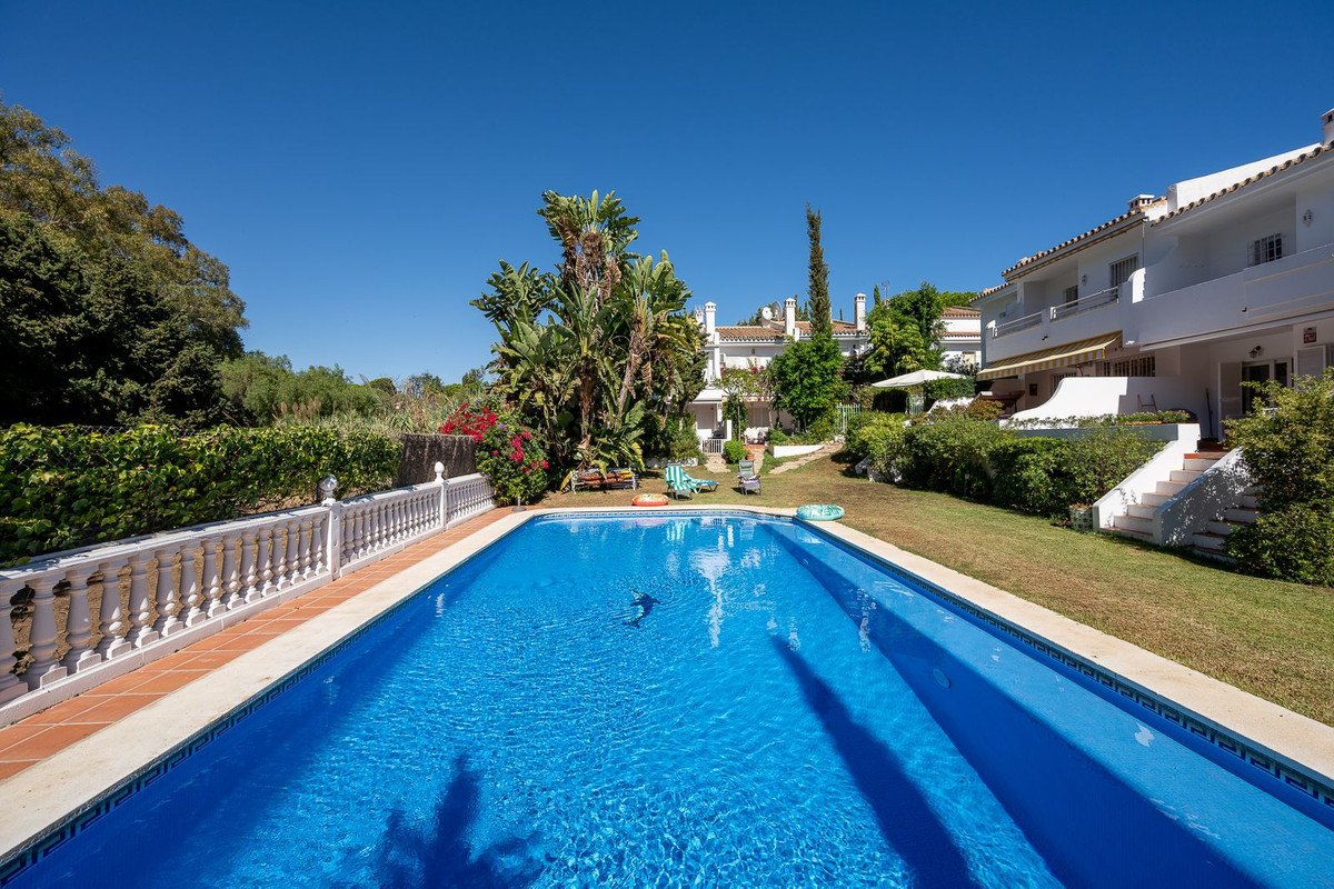 A bright blue swimming pool in a garden, with white villas and lush greenery under a clear blue sky.