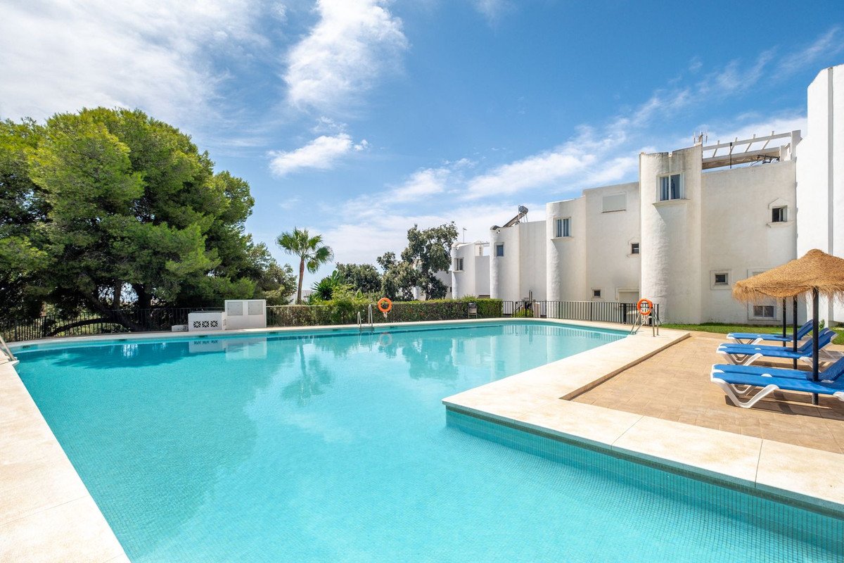 A large, clear blue swimming pool with sun loungers and a thatched umbrella next to white resort buildings under a cloudy sky.