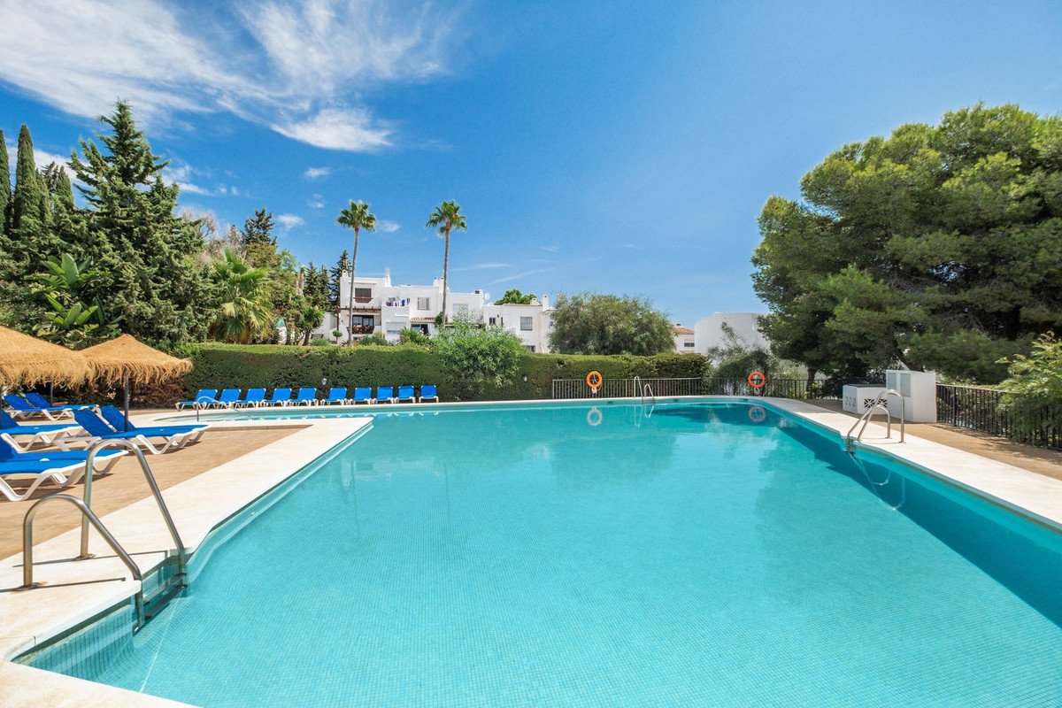 A bright, sunny day at a resort swimming pool with blue lounge chairs and umbrellas, surrounded by greenery and white buildings.