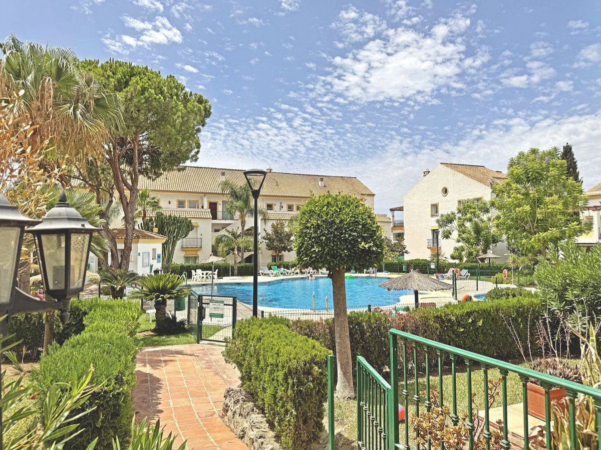 Apartment complex with a blue swimming pool, lush greenery, palm trees, and outdoor seating under a cloudy sky.