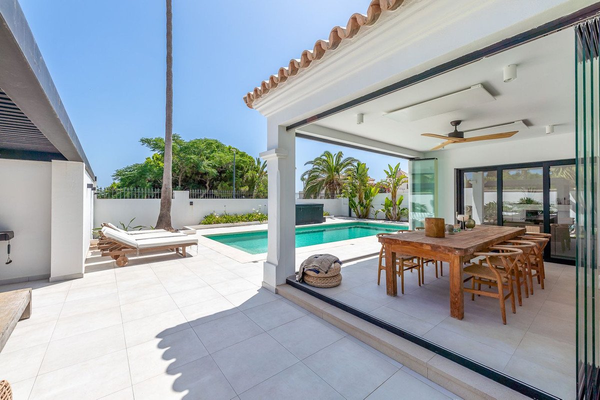 Covered patio with dining table overlooking a swimming pool and sun loungers, surrounded by lush greenery.