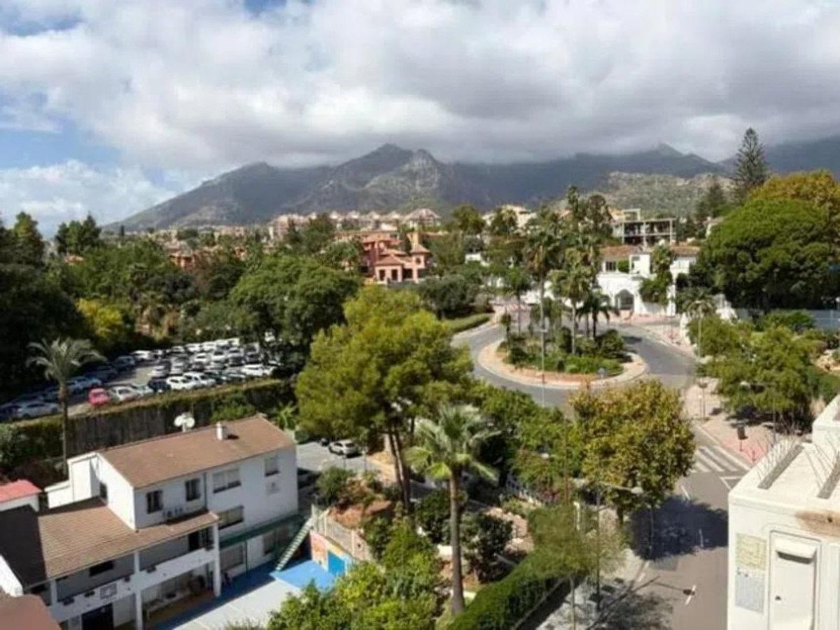 A scenic view of mountains, a street with a roundabout, trees, and buildings under a cloudy sky.