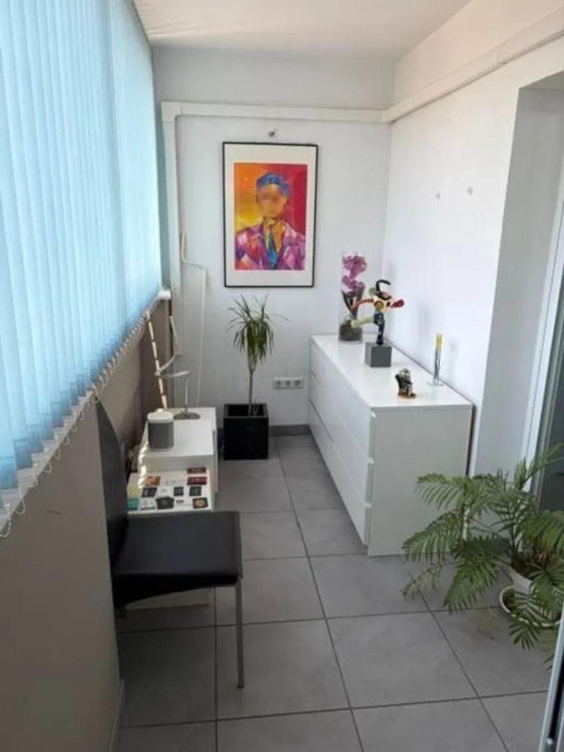 A narrow balcony with a white dresser, abstract portrait art, a chair, and several potted plants.