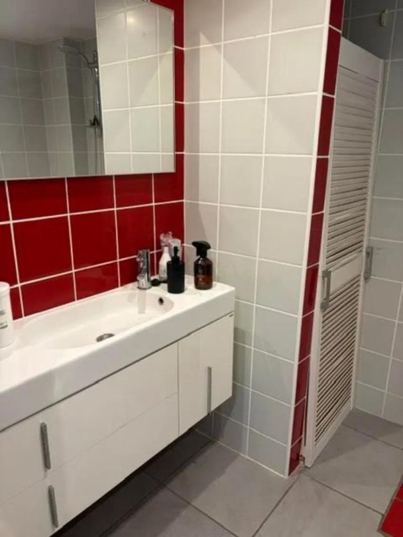 Modern bathroom with a white vanity, red tiled backsplash, and a louvered door. Gray tiled walls and floor.