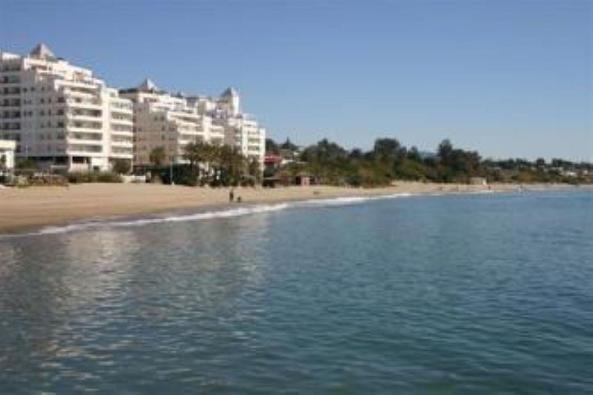 A large white resort building complex overlooking a sandy beach and the blue sea under a clear sky.