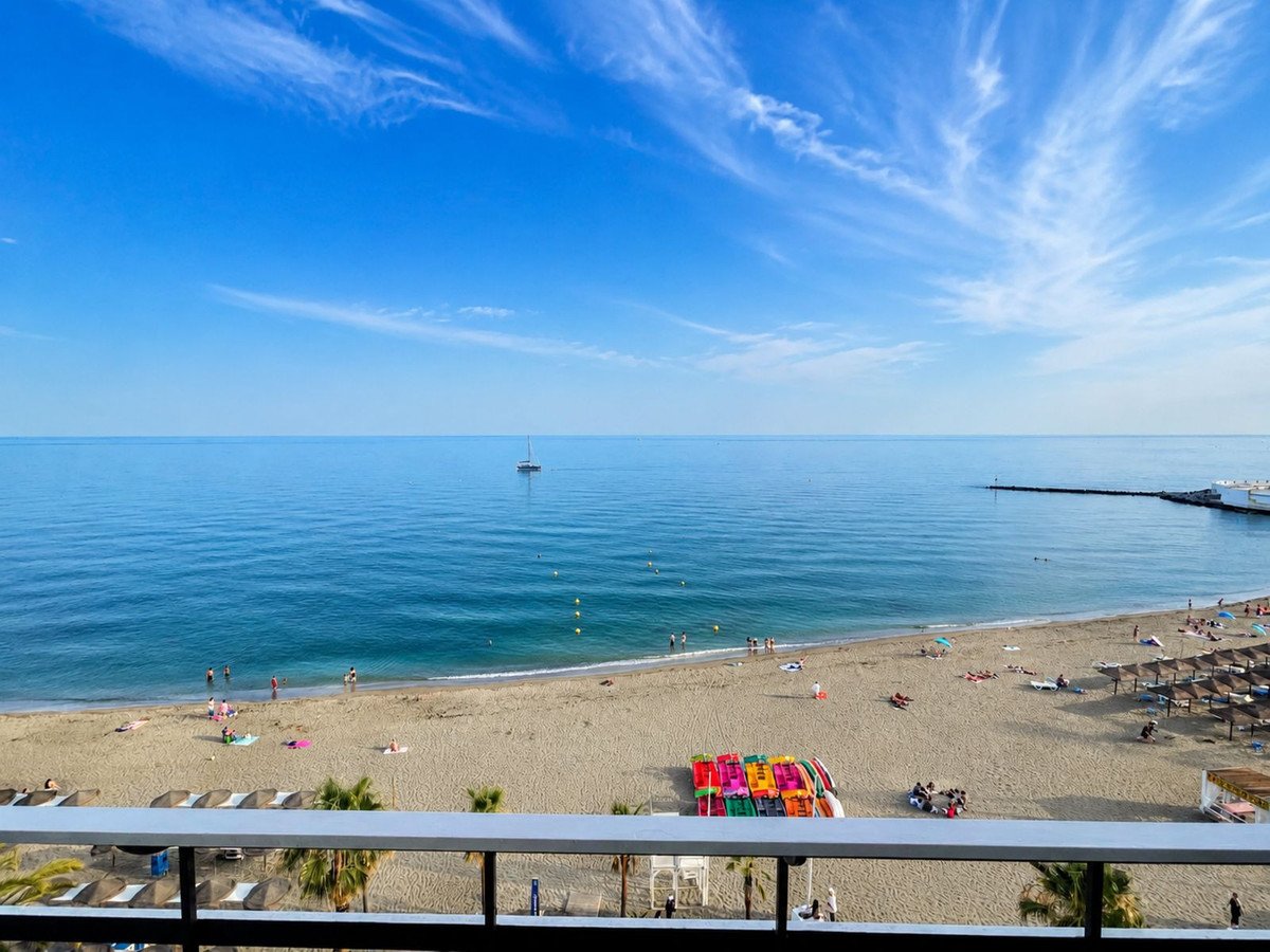View from a balcony overlooking a sandy beach, the blue sea with a sailboat, and people enjoying the day.