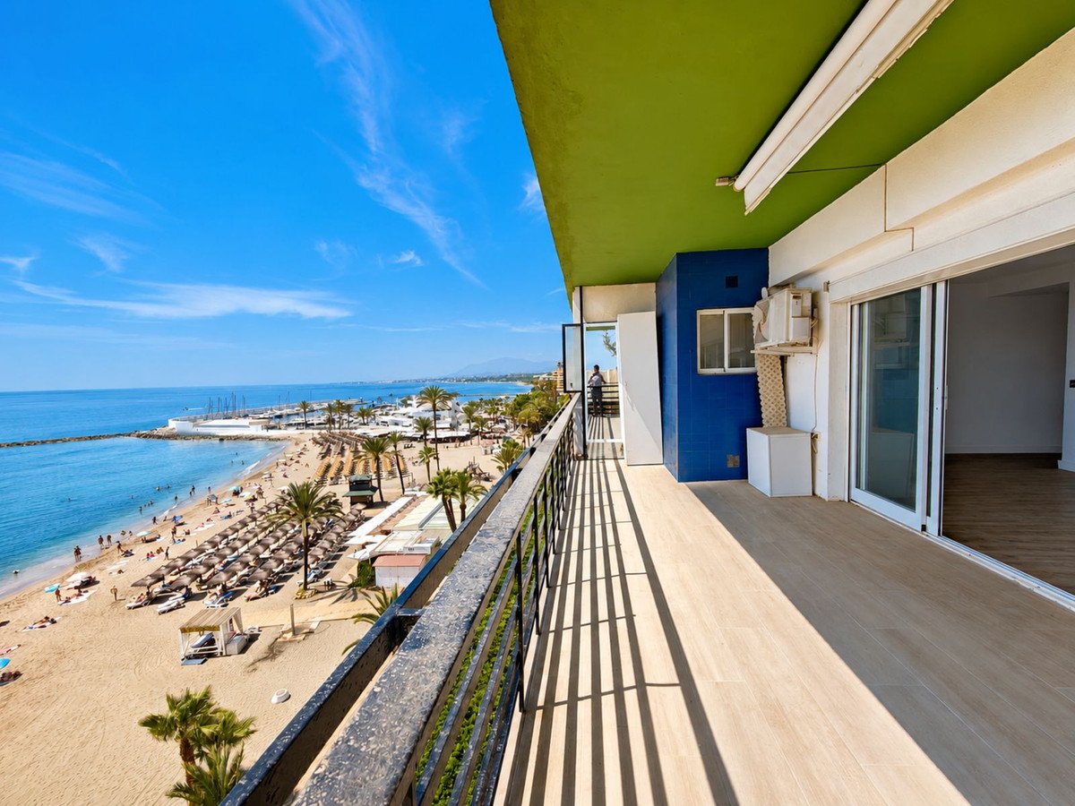 Spacious balcony with a stunning panoramic view of a sandy beach, the blue sea, and a marina on a sunny day.