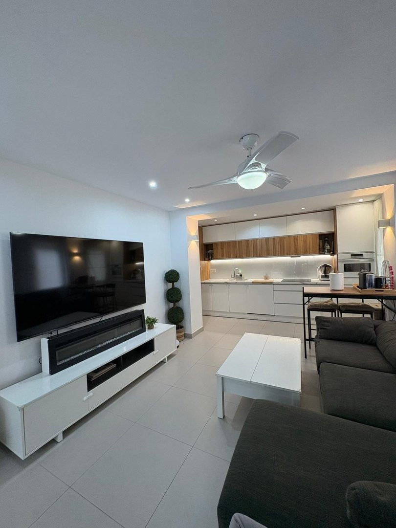 Modern living room with open plan kitchenette, a large TV, a ceiling fan, and a comfortable sofa.