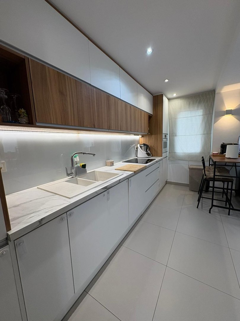 A modern kitchen features white cabinets, marble countertops, a double sink, and wooden upper cabinets. There is a small dining area visible.