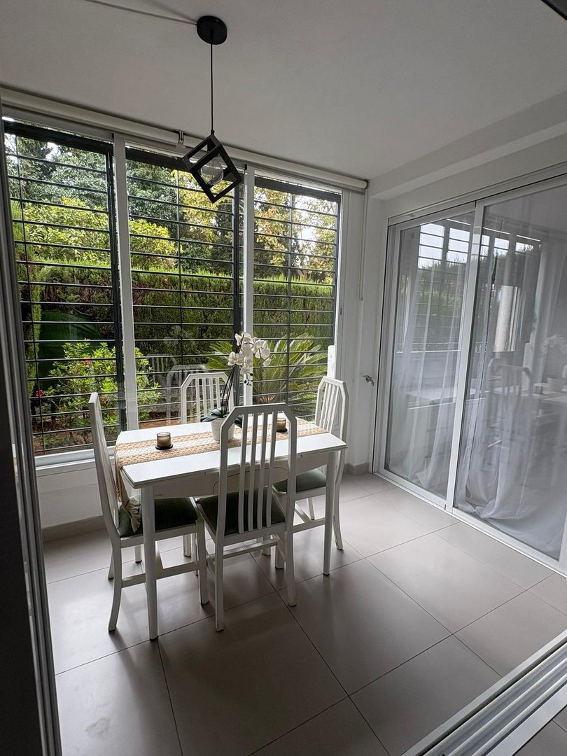 A dining area with white table and chairs, featuring a garden view and abundant natural light from large windows.