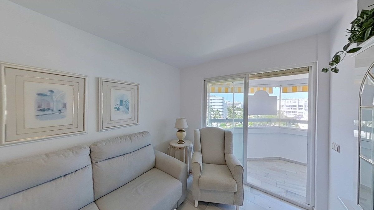 Light living room with sofa, armchair, and balcony view of buildings and trees.