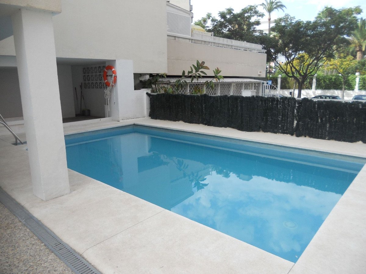 Rectangular swimming pool with clear blue water, surrounded by white concrete and black privacy fence, near a building with a lifebuoy.
