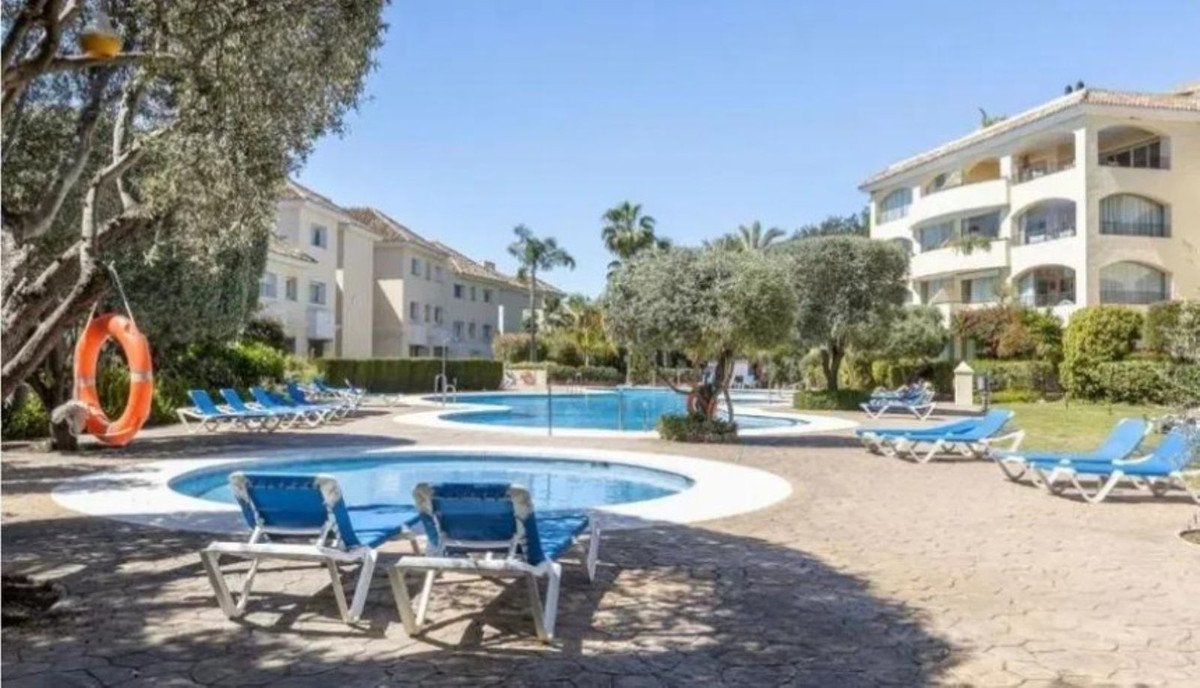 A sun-drenched swimming pool complex with multiple pools, loungers, and apartment buildings in the background.