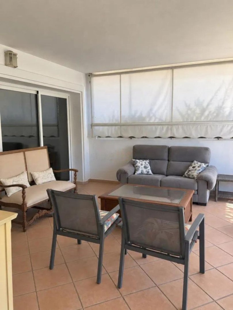 A covered terrace seating area with a sofa, armchairs, and a coffee table, featuring tiled flooring and large sliding doors.