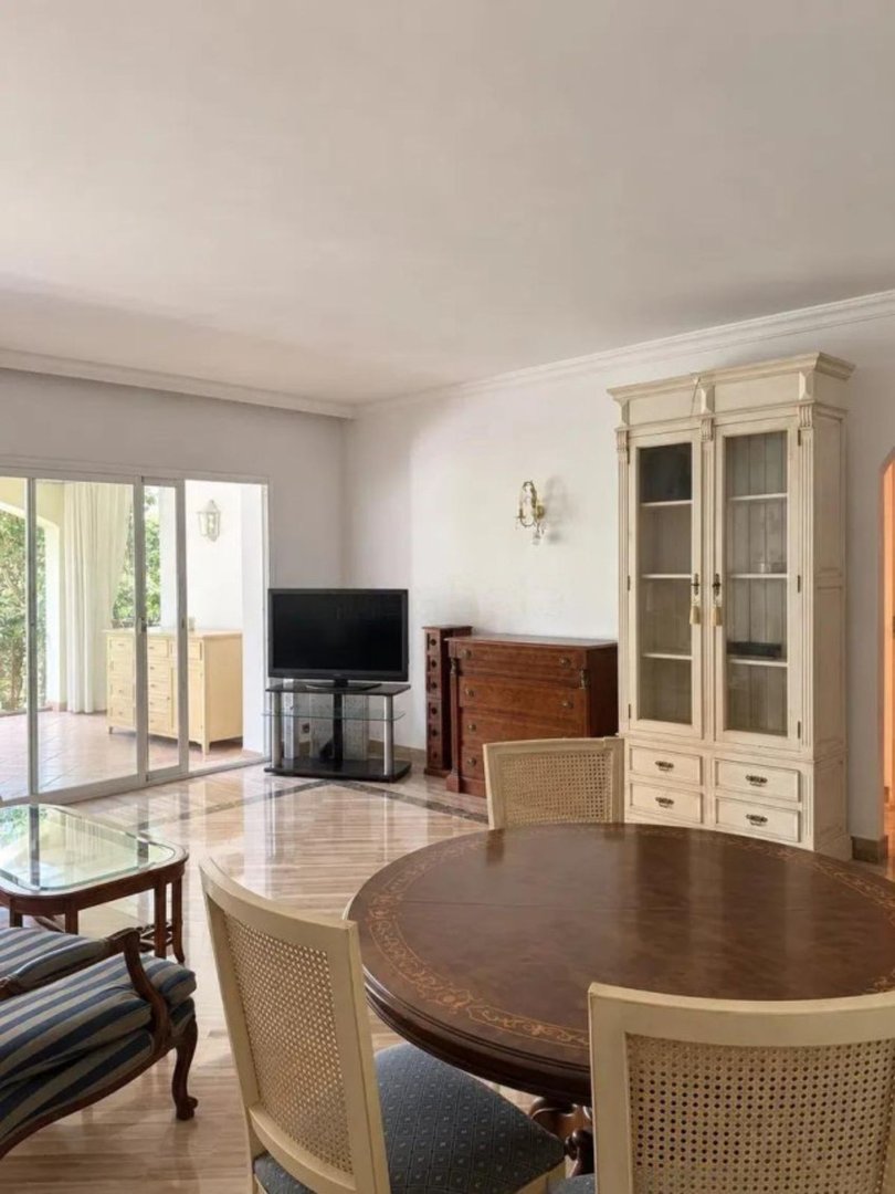 A dining and living area with a large oval table, chairs, a TV, and glass doors leading outside.