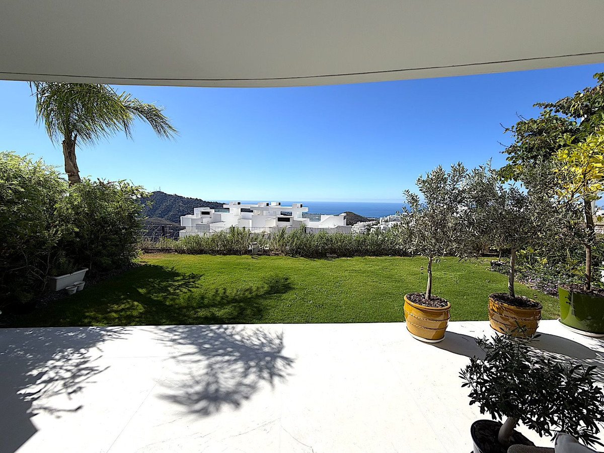 Covered patio with lush green lawn, potted trees, and panoramic ocean and mountain views under a clear blue sky.