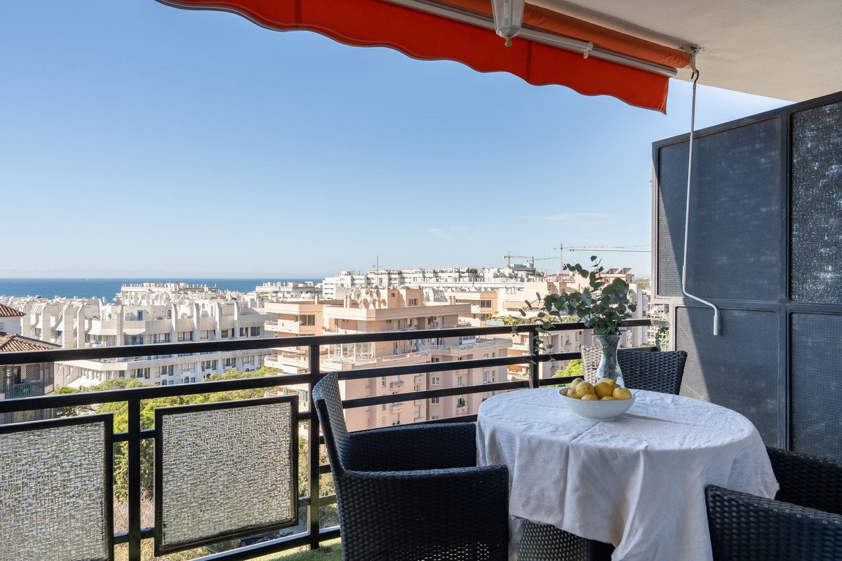 A balcony with a table, chairs, and a view of the sea and cityscape under a sunny blue sky.