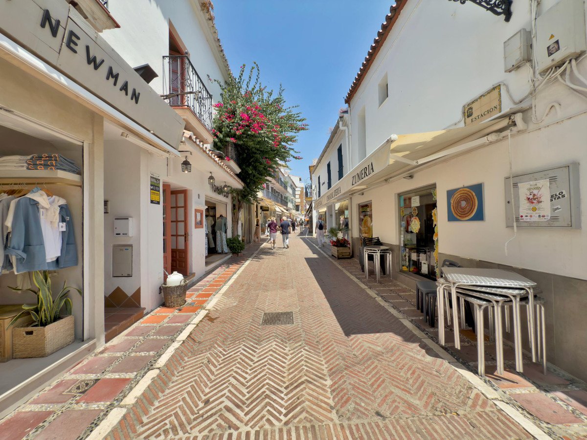 A sunny, narrow street with white buildings, shops, and a herringbone brick walkway.