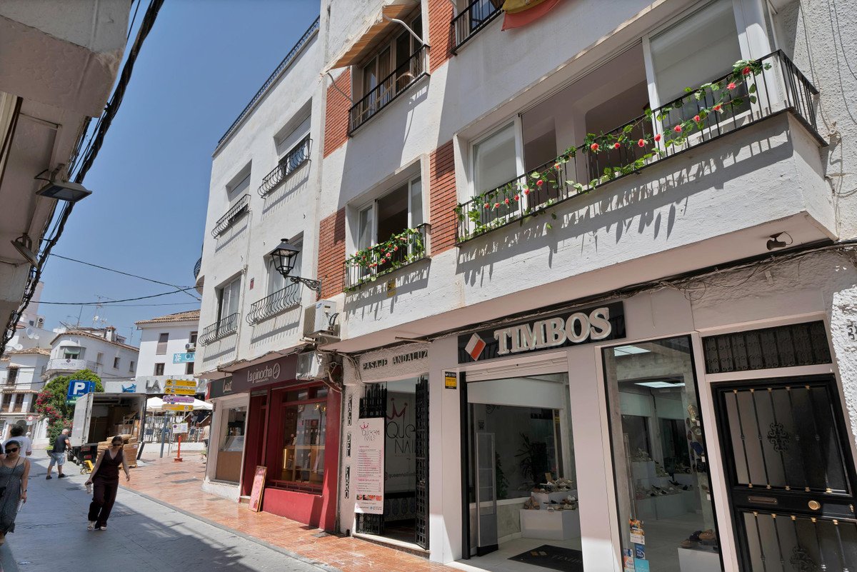A street view of a white building with balconies and shops, including a "TIMBOS" store, under a clear blue sky.