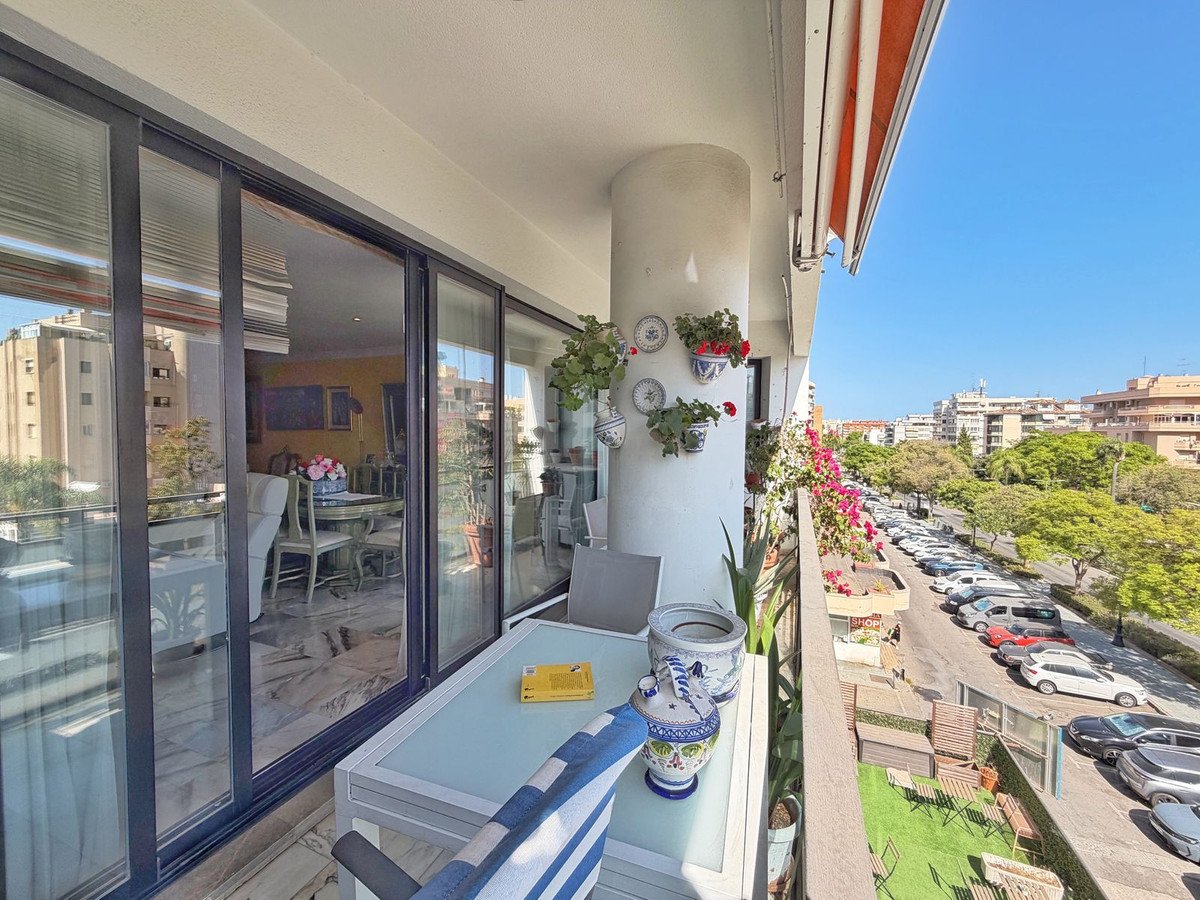 Balcony with potted plants and furniture overlooking a street with parked cars and buildings.