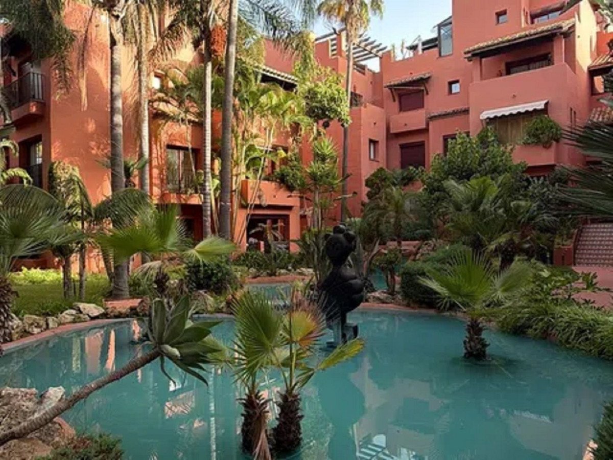A terracotta-colored apartment building with balconies surrounded by lush tropical gardens and a turquoise swimming pool.