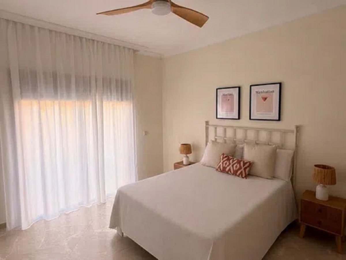 A bedroom featuring a double bed, bedside tables with lamps, and two framed prints on the wall, with large white curtains covering a window.
