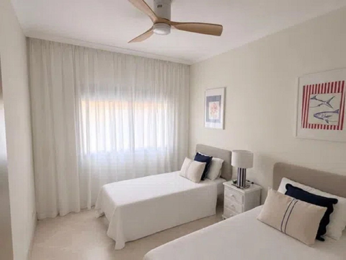 A bright bedroom with two twin beds, white curtains, and a ceiling fan. Decorated with nautical art.