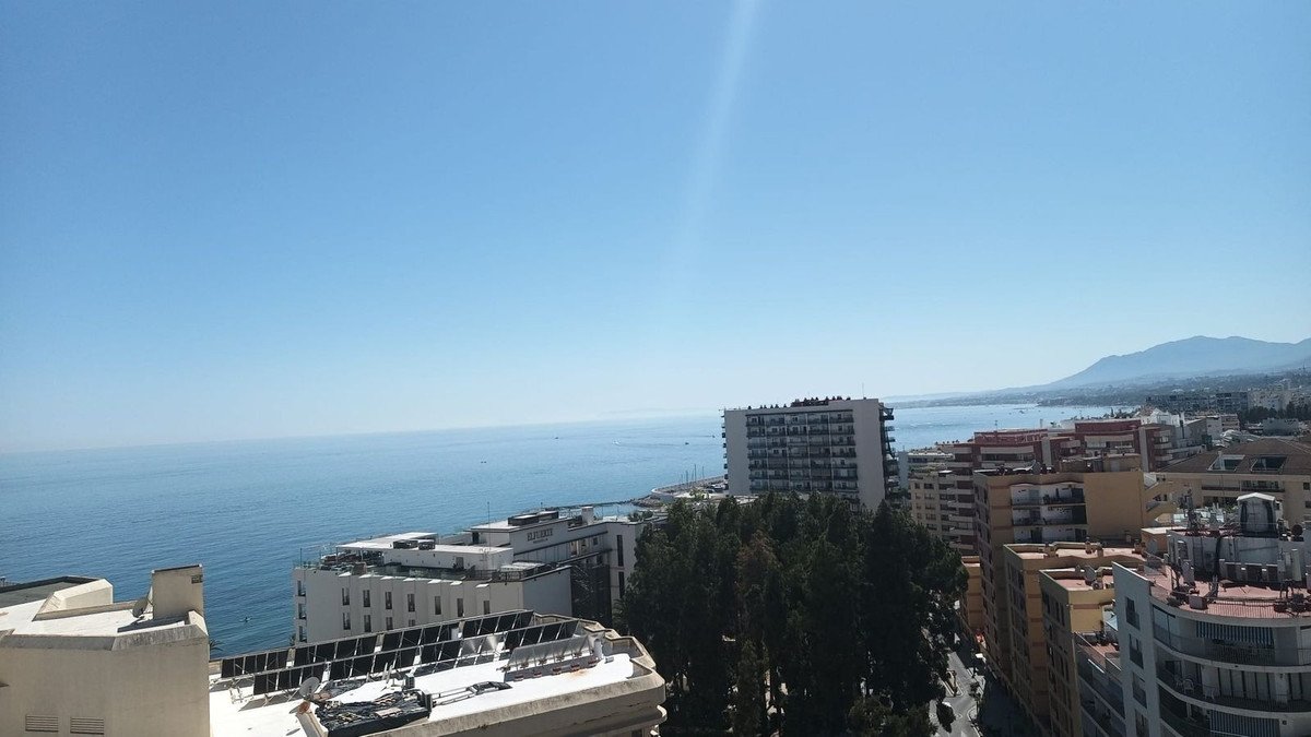 Sea view from balcony, modern buildings, clear blue sky, coastal cityscape.